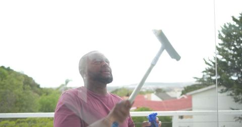 African man cleaning glass balcony panel with long squeegee and blue spray bottle outdoors