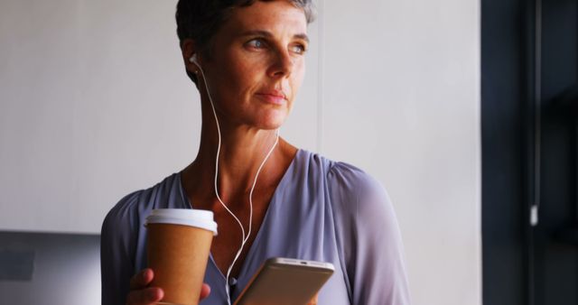 A mature woman in business casual attire is holding a coffee cup and smartphone while wearing headphones. She appears to be taking a break, relaxing and listening to music. This could be used in contexts related to professional life, work-life balance, mid-career professionals, and modern office environments.