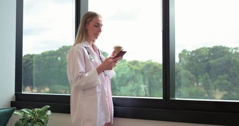 Female doctor holding coffee and checking smartphone by window with stethoscope and plant