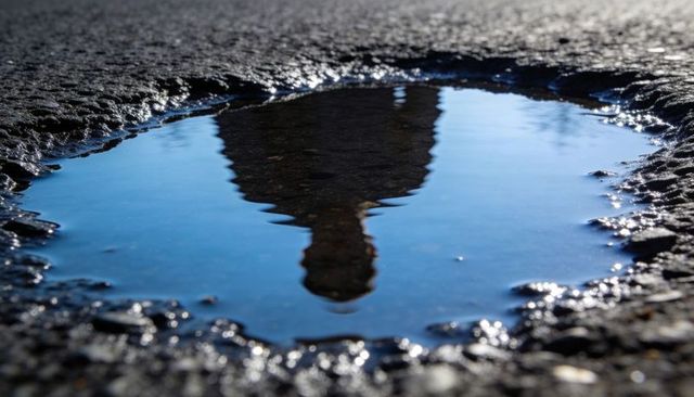 Reflecting puddle showing person silhouette on rough asphalt with blue sky