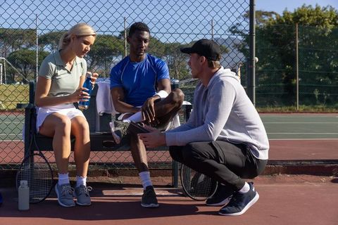 Tennis team huddle with coach using tablet on court bench during outdoor training session