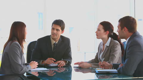 This video depicts four business professionals engaging in a productive meeting. They are seated around a glass table, taking notes and discussing strategy. This scenario can be used for demonstrating teamwork, corporate concepts or illustrating communication in professional settings across corporate publications, presentation slides, and training materials.