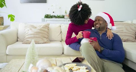 Couple sharing cozy holiday coffee on sofa wearing Santa hat and festive sweaters