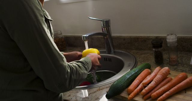 A person is washing yellow bell pepper under running water in kitchen sink. Carrots and cucumber are on the cutting board beside the sink. Suitable for use in blogs, articles, and advertisements related to health, cooking tutorials, and home lifestyle.