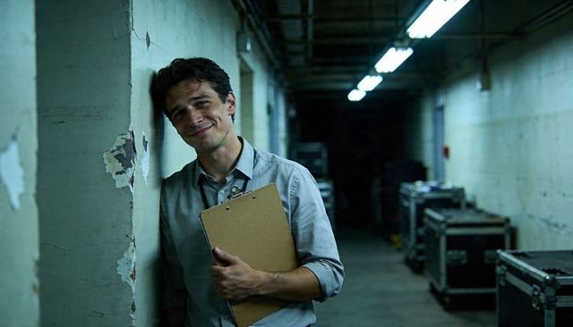Backstage technician leaning on wall holding clipboard in dim backstage corridor