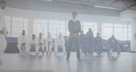 Standing official officiating martial arts match on mat with competitors seated in gi