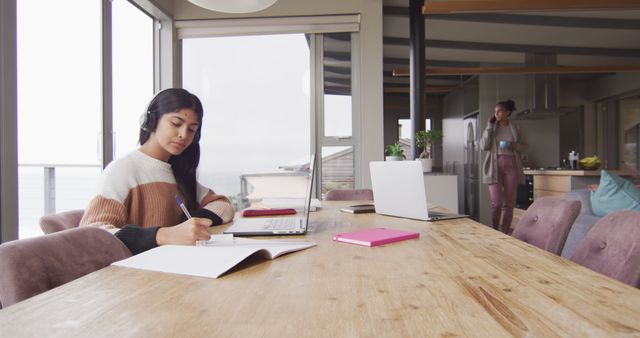 This image captures a young woman working or studying at home, focusing on her studies while using headphones. The modern home interior creates a calm and productive atmosphere, ideal for concepts related to education, remote work, or home office setup. Useful for promoting online learning, digital education platforms, or work-from-home products.