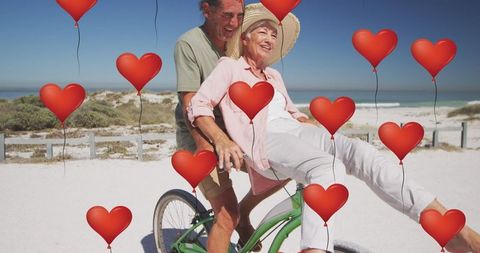 Joyful senior couple riding bicycle on beach with heart icons