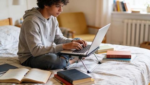 Teen Studying on Bed Using Laptop on Portable Folding Stand with Textbooks for Remote Learning