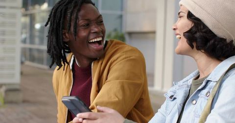 Laughing diverse friends sharing smartphone on urban bench near modern architecture