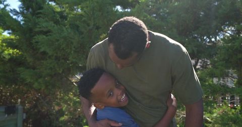 African american father hugging son and smiling in sunlit backyard garden, family bond
