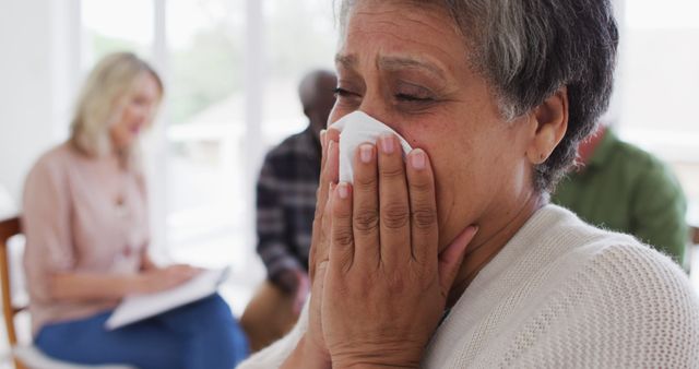Elderly woman crying and using a tissue while attending a support group therapy session. The emotional moment captures her deep feelings, and others in the background provide a supportive environment. Suitable for mental health, counseling, therapy, senior care, and emotional support themes, as well as illustrating concepts of grieving and coping mechanisms. Ideal for blog illustrations, mental health websites, support group promotional material, and educational content regarding mental well-being.