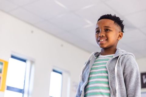Smiling african american boy standing in bright classroom wearing gray hoodie striped tee