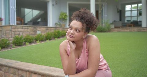 Contemplative woman in pink activewear sitting on stone wall in modern backyard