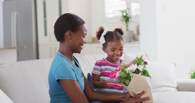 Happy african american mother and daughter sitting on sofa and smelling flowers. staying at home in self isolation during quarantine lockdown.