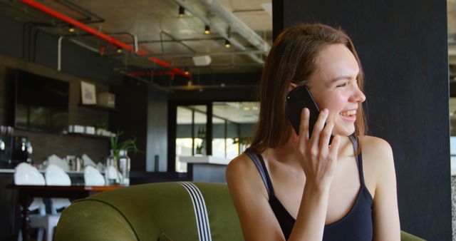 Young woman smiling and laughing while having a phone conversation. Musturds10 setting in a modern living room featuring a green couch, industrial design elements and natural light. Ideal for stock photos about lifestyle, communication, relaxation, and modern interiors.