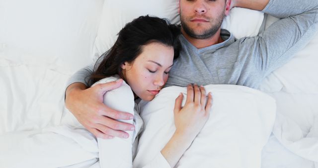 Young couple lying in bed, peacefully sleeping while cuddling under white sheets. Ideal for concepts related to romantic relationships, intimacy, comfort, and relaxation. Suitable for usage in lifestyle blogs, relationship advice articles, sleep-related content, and advertisements for bedding or sleep products.