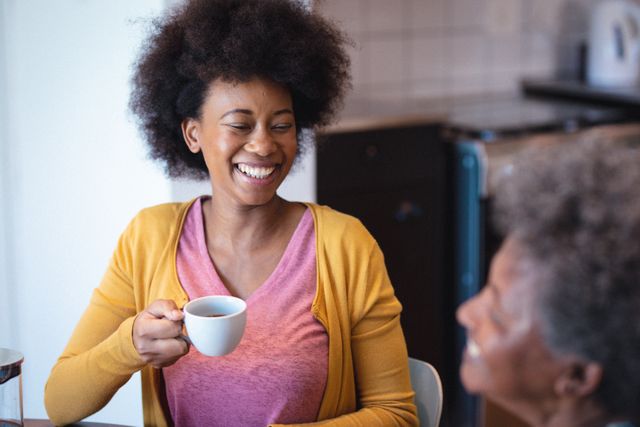 African American female physiotherapist holding a coffee cup and smiling while sitting with a senior woman at home. This image can be used for promoting senior homecare services, healthcare advertisements, or lifestyle blogs focusing on elderly care and companionship.