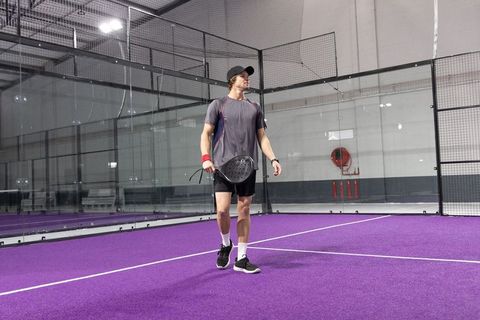 Male padel player walking on vibrant purple indoor court holding paddle and wearing cap