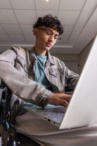Teenage male typing on laptop in wheelchair studying in minimalist study room with glasses