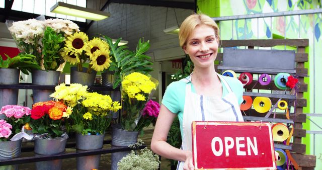 Florist standing at entrance of a flower shop, smiling while holding an 'Open' sign. Various colorful flower arrangements on display, creating a vibrant and inviting atmosphere. Suitable for illustrating themes of small business, reopening, welcoming customers, seasonal sales, or promoting flower shops.