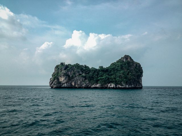Image of a solitary, green-covered island amidst calm, blue ocean water under a cloudy sky. Ideal for travel brochures, adventure blogs, and nature photography collections.