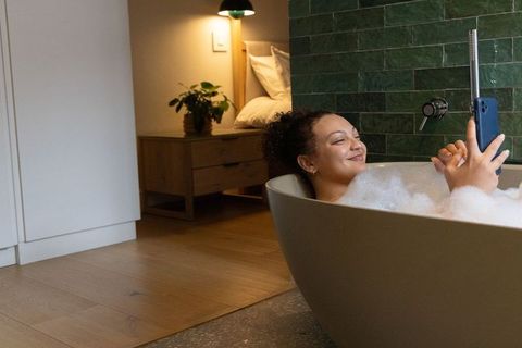 Relaxing african american woman taking selfie in bubble bath in modern home bathroom