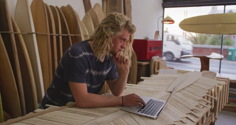 Surfboard Maker Contemplating Design on Laptop in Workshop