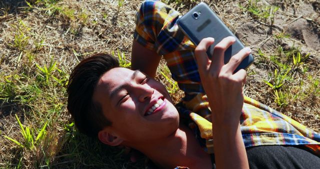 Young man lying on grass and smiling while using smartphone. Perfect for topics related to relaxation, technology usage, youth lifestyle, outdoor leisure activities, and communication. High relevance for social media posts, advertisements, blog articles on relaxation, as well as marketing campaigns targeting young adults.