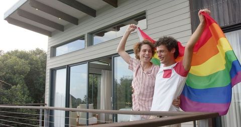 Joyful Gay Couple Embracing on Balcony with Vibrant LGBT Flag