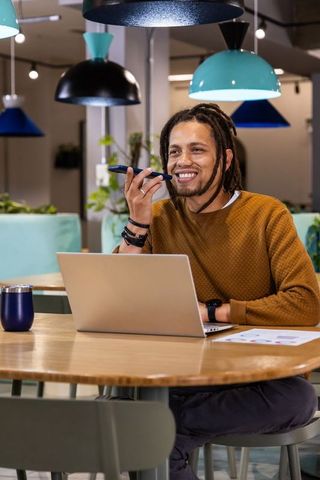 African American man speaking into smartphone while typing on laptop in coworking cafe