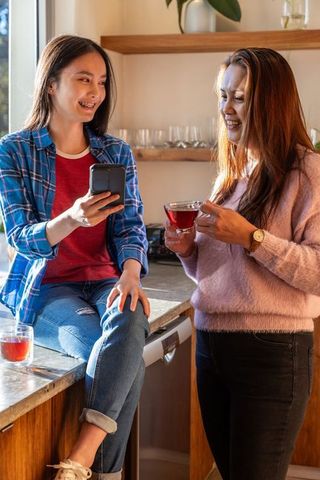 Asian Mother and Daughter Chatting in Cozy Kitchen Holding Smartphone and Tea