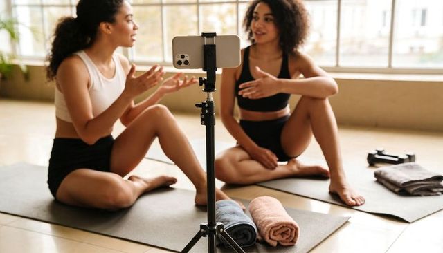 Two fitness partners filming yoga tutorial on smartphone tripod while chatting