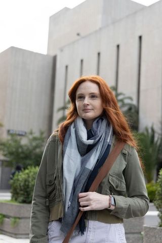 Red-haired woman standing in urban plaza wearing scarf, utility jacket and crossbody bag