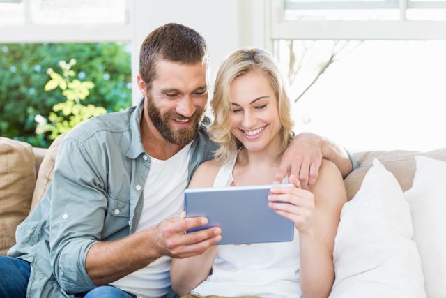 Happy couple sitting on a sofa in a bright living room, using a digital tablet together. They are smiling and appear relaxed, enjoying their time at home. This image is ideal for use in advertisements, blogs, and articles related to technology, home life, family bonding, and modern lifestyles.