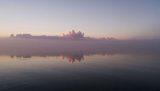 Sunlit cumulus bank reflecting on calm sea at dawn with pastel mist and mirror horizon