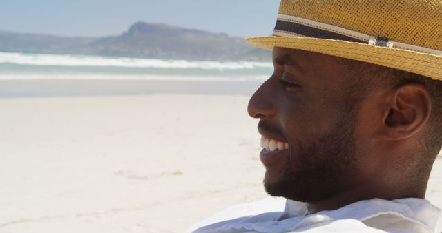 Man wearing straw hat and smiling while enjoying a sunny day at the beach. Ideal for promoting summer vacations, trips, and beach leisure activities. Could also be used in advertisements focusing on relaxation, happiness, and outdoor enjoyment.