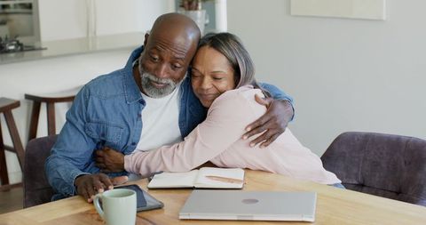 Mature Black couple embracing while sharing laptop and tablet at cozy kitchen table