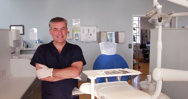 Smiling dentist standing in a dental clinic with arms cross. He is surrounded by various dental equipment, including a dental chair and instruments. This image is perfect for websites, brochures, and advertisements related to dental care, oral hygiene, healthcare professionals, and medical institutions.