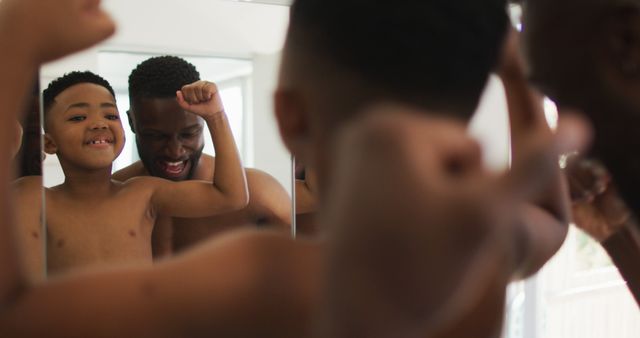African american father and son flexing their muscles in mirror together. staying at home in self isolation during quarantine lockdown.