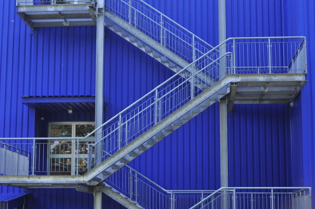 Shows a modern metal staircase attached to the vibrant blue exterior of an industrial building. Represents contemporary architectural elements, urban settings, and industrial designs. Ideal for illustrating modern architecture, urban planning, safety compliance visuals, and geometric pattern-themed projects.