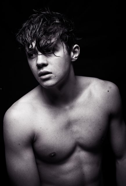 A young man with wet hair posed shirtless in dramatic lighting, shot in black and white. This can serve as an artistic portrait or be utilized in promotional material for fitness, wellness, and grooming products. Additionally perfect for editorial content requiring a strong, edgy visual.