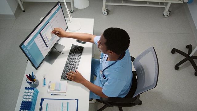 Male clinician in scrubs reaching touchscreen monitor while typing at clinic workstation