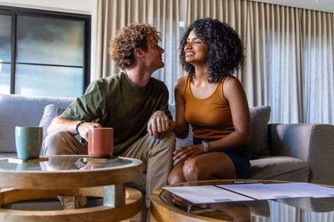 Mixed-race couple holding hands on modern sofa, sharing coffee and reviewing documents