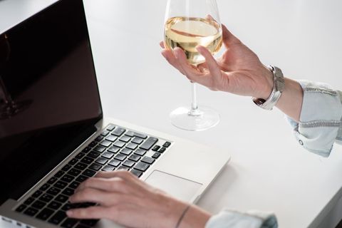 Typing on laptop while holding white wine glass for relaxed remote work and multitasking