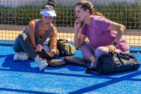 Two women relaxing on bright blue paddle court checking smartphone between matches