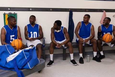 African american basketball team sitting on bench in locker room wearing blue jerseys