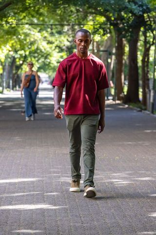 African american man walking on sunlit tree-lined path wearing burgundy shirt, olive pants