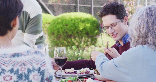 Group of friends having a good time around a table enjoying a healthy meal with red wine in a garden setting. Perfect for use in advertising recipes, outdoor dining experiences, social gatherings, friendship, and lifestyle blogs.