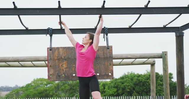 Determined caucasian woman in pink t shirt hanging from monkey bars on bootcamp training course. Female fitness, challenge and healthy lifestyle.